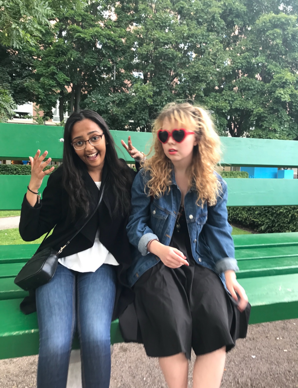 Two friends on a green park bench, one wearing red heart-shaped sunglasses
