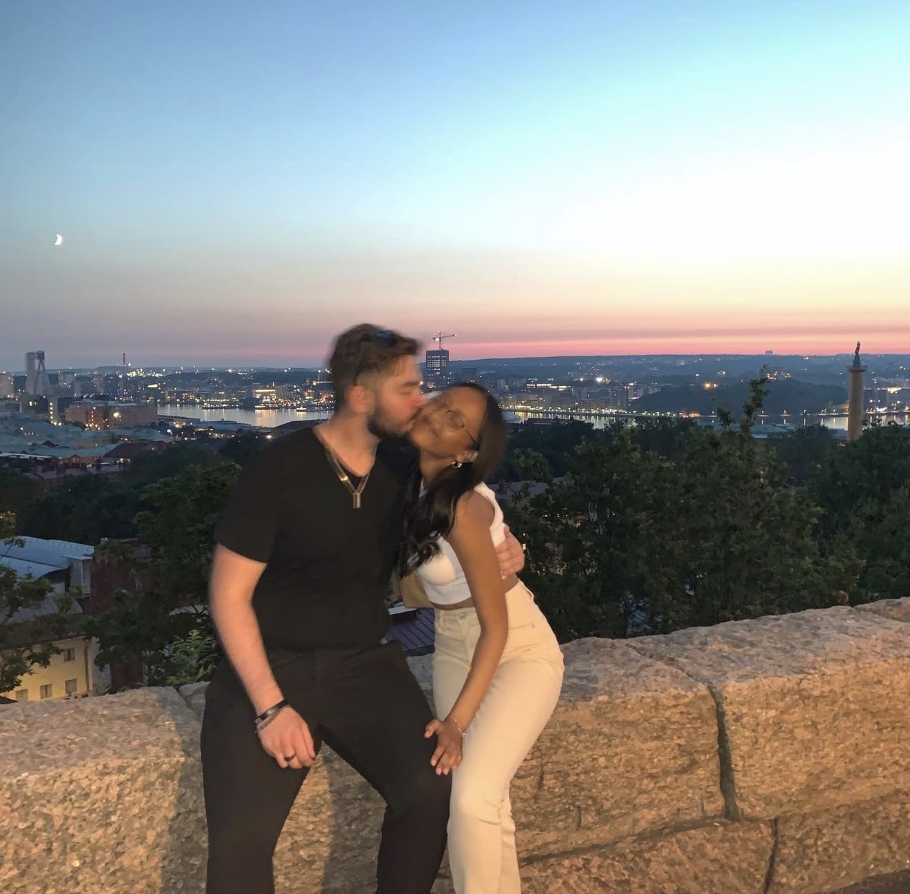 Two friends sharing a kiss on the cheek with a pink sunset and city skyline behind them