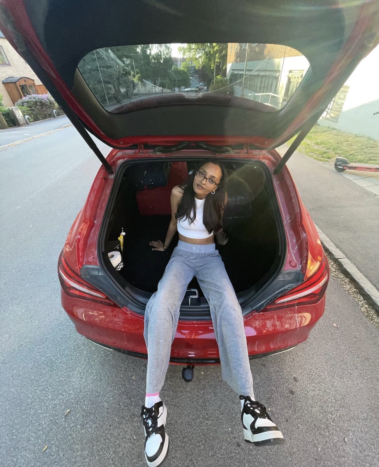 A woman in glasses sitting in the open boot of a parked red car