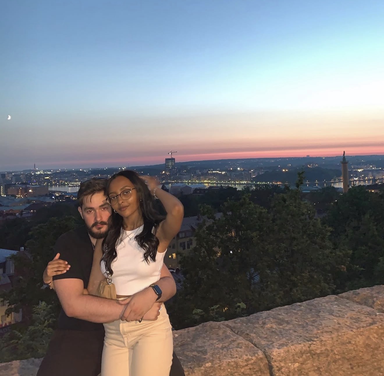 Two friends together beside a stone wall overlooking a city at sunset