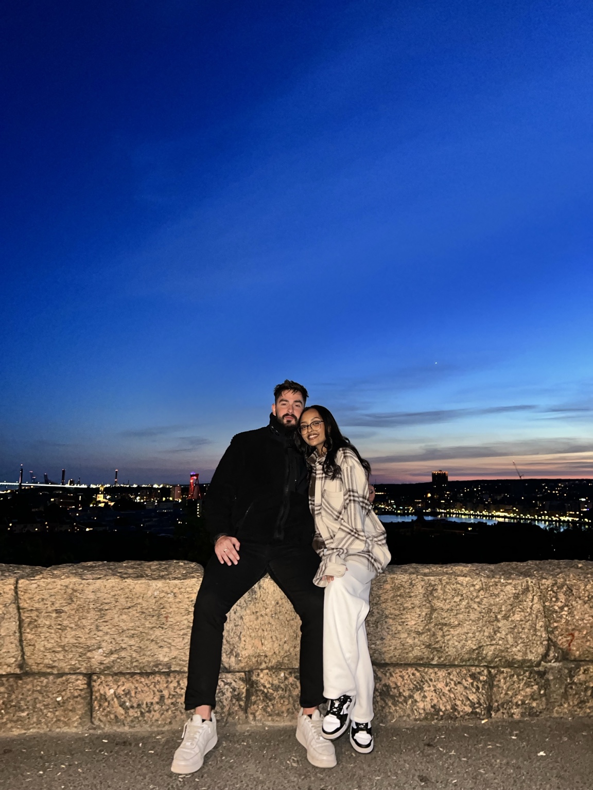 Two friends leaning against a stone wall above a city lit up at dusk